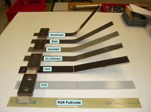 Seven material samples after 3-point bending drop test — metals permanently deformed, wood broken, PVC damaged, PUR pultruded profile intact